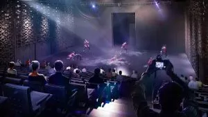 Un groupe de danseurs se produit sur scène, éclairé par des projecteurs. Les spectateurs observent depuis des sièges en gradins, tandis qu'un individu prend une photo depuis l'arrière. La salle est sombre.