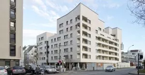Bâtiment moderne beige à plusieurs étages avec balcons, situé en bordure de rue. En bas, voitures garées et panneau de signalisation. Ciel dégagé et quelques arbres autour.