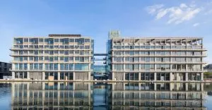 Deux immeubles modernes, reliés par une passerelle en verre, se dressent côte à côte. Ils se reflètent dans un plan d'eau calme, sous un ciel dégagé.