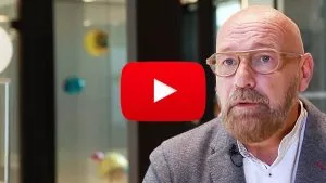 Un homme barbu avec des lunettes parle dans un environnement moderne et lumineux. Le logo de YouTube est superposé sur l'image, indiquant un contenu vidéo.