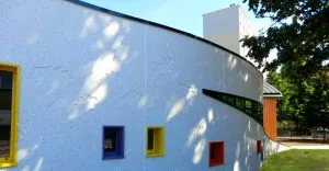 Bâtiment moderne aux murs courbes blancs avec incrustations, comportant des fenêtres colorées jaunes, bleues et orange. Contexte arboré, avec ombres des arbres projetées sur la façade sous un ciel ensoleillé.