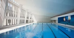Piscine intérieure moderne avec une large baie vitrée laissant entrer la lumière naturelle. Un plongeon crée des éclaboussures dans l'eau. Un homme se dirige vers les gradins bleus.