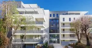 Bâtiment moderne avec façades blanches et balcons ornés, entouré d'arbres au feuillage léger. Le ciel est dégagé et l'environnement semble urbain et calme. Aucun texte présent.