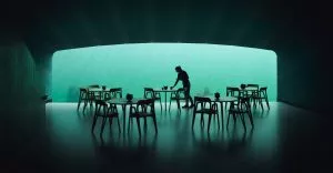 Une personne nettoie une table dans un restaurant sous-marin, entouré de nombreuses tables et chaises. À travers une grande vitre, on voit la mer éclairée d'une lumière turquoise.