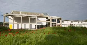 Bâtiment moderne, en partie vitré, avec de grandes poutres en béton, se tient dans un champ de coquelicots rouges sous un ciel nuageux. Une porte jaune est visible.
