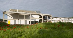 Bâtiment moderne, en partie vitré, avec de grandes poutres en béton, se tient dans un champ de coquelicots rouges sous un ciel nuageux. Une porte jaune est visible.