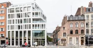 Un immeuble moderne blanc, rectangulaire avec de grandes fenêtres, se dresse à côté de bâtiments anciens en brique. Deux panneaux STOP rouges sont visibles devant, sur une rue urbaine.