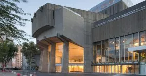 Bâtiment moderne aux larges fenêtres vitrées éclairées, montrant une façade en béton angulaire sur pilastres, situé dans un quartier urbain avec arbres et autres bâtiments autour. Environnement calme et moderne.