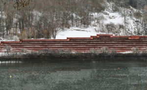 Structure rouge en terrasses s'étend le long d'un lac calme, bordée par des arbres enneigés sur une colline. Aucune inscription n'est présente dans l'image.