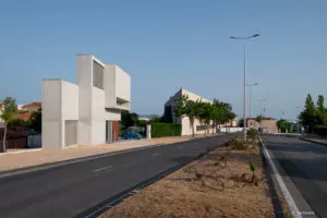 Un bâtiment moderne en béton géométrique se tient immobile près d'une route. Des arbres bordent la rue, et une voiture bleue est garée devant le bâtiment.