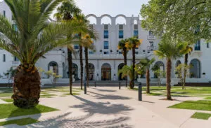 Bâtiment blanc avec arcs en façade, entouré de palmiers élancés. L'entrée vitrée est bordée d'arbres et d'une pelouse, offrant une atmosphère lumineuse et accueillante.