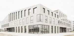 Bâtiment moderne aux lignes géométriques, grandes fenêtres en verre, situé dans un environnement urbain dégagé. Le sol est pavé devant, sans inscription visible.