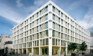 Bâtiment moderne avec façades blanches et vitrées, situé à l'angle d'une rue urbaine, entouré de trottoirs, de lampadaires et de quelques arbres. Le ciel est légèrement nuageux.
