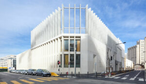 Bâtiment moderne blanc avec de grandes fenêtres verticales situé à un carrefour urbain. Voitures stationnées le long de la rue, ciel bleu en arrière-plan. Environnement urbain avec signalisation routière.