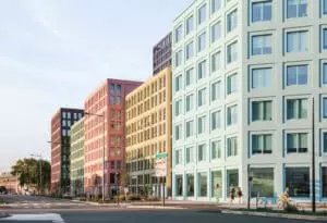 Immeubles colorés alignés le long d'une rue, chacun d'une teinte distincte: vert, rose, beige. Passants marchant ou à bicyclette. Climat clair, végétation peu abondante. Panneaux de signalisation présents.