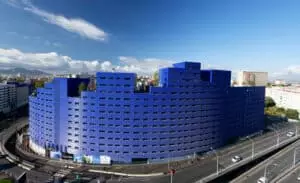 Bâtiment bleu incurvé entouré de routes, situé sur une avenue urbaine avec quelques voitures passant à proximité. Des immeubles et un ciel bleu complètent le paysage en toile de fond.
