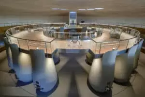 Une salle circulaire moderne avec de grandes cuves métalliques grises autour d'une plateforme centrale en bois, protégée par des barrières en verre. Environnement industriel épuré avec éclairage naturel.