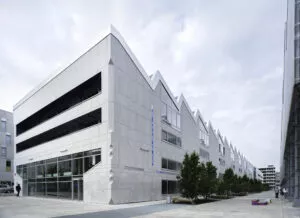 Bâtiment moderne aux lignes géométriques avec des fenêtres rectangulaires, situé long d'une allée arborée pavée. Des personnes marchent à côté; le ciel est nuageux. Une enseigne indique "ÉCOLE". Texte visible : "ÉCOLE D'ARCHITECTURE", "BAT B / NORD - ENTRÉE A", "ENTRÉE - LIBRAIRE".
