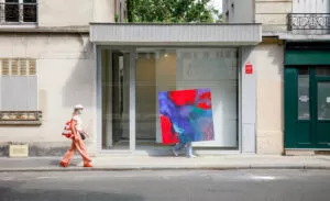 Une grande peinture abstraite colorée, principalement rouge et violet, est transportée par une personne au bord d'un trottoir devant une galerie. Une personne passe en tenue beige et chapeau.
