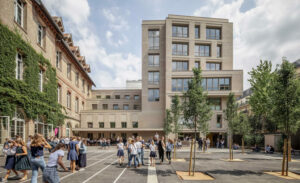 Une cour d'école animée, avec des élèves jouant et discutant, est entourée par des bâtiments en briques recouverts partiellement de lierre. Des lignes blanches marquent le sol, et de jeunes arbres sont plantés.