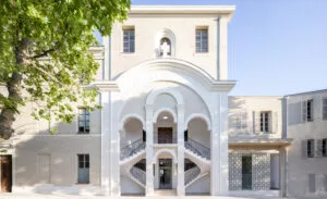 Un bâtiment beige à trois niveaux avec des arches blanches, entouré d'arbres et de ciel bleu. L'entrée présente un double escalier symétrique sous un grand arc central.