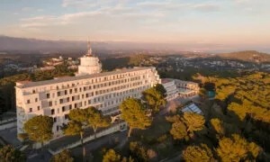 Bâtiment blanc imposant avec antenne sur le toit, se dresse au-dessus d'une forêt de pins verdoyants. En arrière-plan, montagnes et ville sous une lumière de fin de journée.
