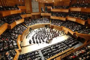 Un orchestre joue sur une scène circulaire en bois, entouré de gradins remplis de spectateurs. L'environnement est une salle de concert moderne et spacieuse.