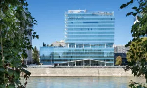 Un bâtiment moderne avec de nombreuses fenêtres en verre réfléchissant se dresse au bord de l'eau. Le nom "KAUFMAN & BROAD" est affiché en haut. Des arbres encadrent la vue.