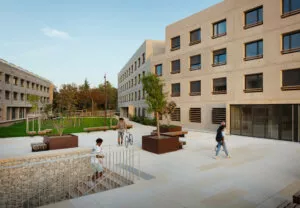 Un espace extérieur moderne avec des bâtiments en béton alentour. Des personnes marchent ou roulent à vélo sur une vaste place pavée, entourée de plantes et d'arbres. Le ciel est clair.