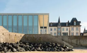 Bâtiment moderne aux grandes fenêtres vitrées se tenant à côté d'une maison traditionnelle avec tourelles, encadré par un mur de pierre. L'ensemble est situé près d'une plage au sable doré.
