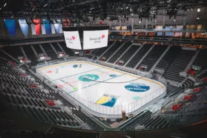 Patinoire située dans une arène vide, entourée de gradins. Des bannières colorées pendent du plafond. Les écrans affichent "Swiss Life Arena". Environnement moderne et spacieux.