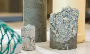 Cylindres de béton renforcés de fibres colorées sont disposés sur une surface blanche. L'un est cassé, révélant des gravats et fibres à l'intérieur. L'arrière-plan est flou.