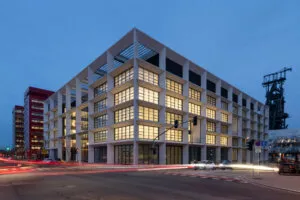 Bâtiment moderne illuminé au crépuscule, angle de rue avec circulation floue de voitures, situé dans un quartier urbain, environné de structures industrielles et autres bâtiments modernes.
