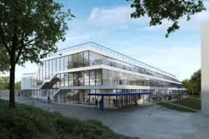 Bâtiment moderne à plusieurs étages avec de grandes baies vitrées, entouré d'arbres et de végétation, des personnes se déplacent à l'extérieur, ciel dégagé en arrière-plan.