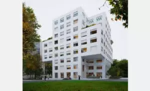 Bâtiment blanc cubique avec de nombreuses fenêtres, se dresse au centre. Les personnes marchent en bas, entourées d'arbres verdoyants. L'atmosphère est moderne et urbaine.