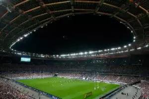 Stade éclairé avec un match de rugby en cours, entouré de spectateurs. Le ciel est sombre, et la lune est visible. Une grande tribune et un écran montrent le jeu.