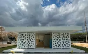 Un petit bâtiment moderne avec des panneaux perforés se dresse immobile sous un ciel nuageux. Il est entouré de structures en bois et de verdure, suggérant un cadre extérieur.