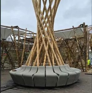 Un banc circulaire gris est disposé autour d'une structure en bambou entrelacée. Le contexte est un espace extérieur en bois, entouré de constructions en bambou et de toiles tendues.