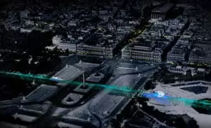 Vue aérienne d'une grande place avec une obélisque centrale, illuminée par des lignes lumineuses bleues et vertes. Le contexte montre des bâtiments environnants détaillés et des rues sombres la nuit.