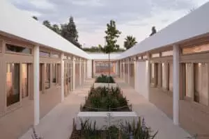 Un corridor extérieur bordé de fenêtres en bois, entourant des plates-bandes végétales centrales. Le ciel est légèrement nuageux, et des arbres apparaissent en arrière-plan.