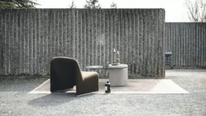 Chaise brune sur un tapis, à côté d'une table basse avec livres et chandeliers. Contexte : mur en béton ondulé, arbres en arrière-plan, surface gravillonnée.