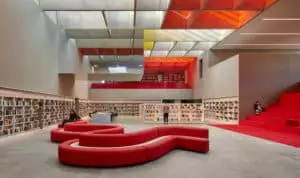 Des canapés rouges forment un espace de détente au centre d'une bibliothèque moderne avec des étagères. Des personnes lisent et explorent les livres. Un escalier rouge conduit à une mezzanine.