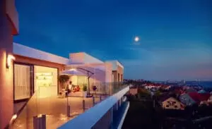 Terrasse moderne éclairée le soir avec parasol et mobilier confortable, deux personnes assises conversant. Vue panoramique sur une ville et maisons sous un ciel crépusculaire, lune visible.