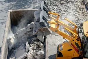 Une pelle mécanique jaune charge des blocs de béton cassés dans une benne, entourée de poussière et de débris sur un chantier de construction en plein air.