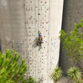 Un grimpeur, équipé d'un casque, escalade un mur d'escalade en béton avec des prises colorées. Autour, des arbres verts entourent la structure, créant un environnement naturel.