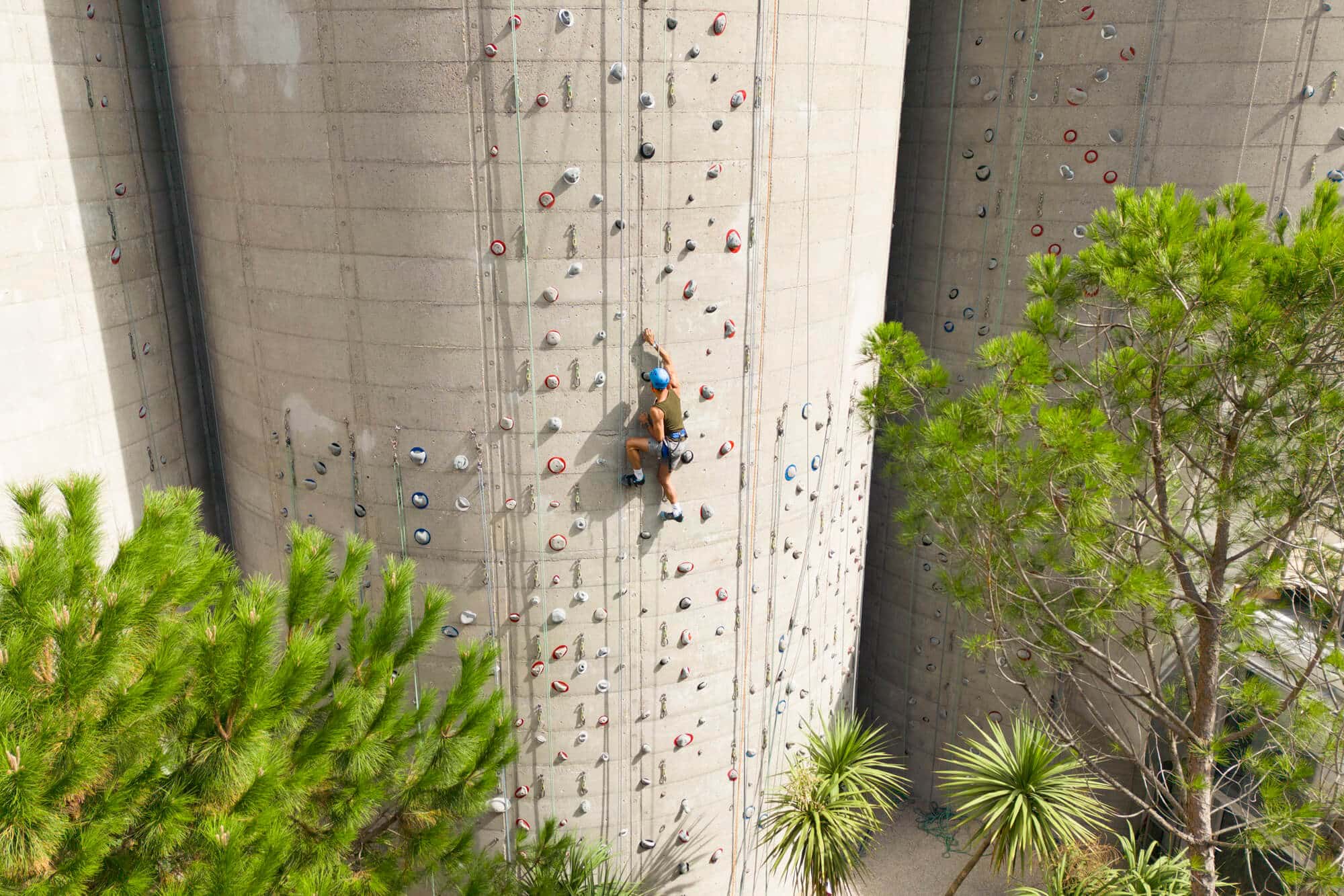 Un grimpeur, équipé d'un casque, escalade un mur d'escalade en béton avec des prises colorées. Autour, des arbres verts entourent la structure, créant un environnement naturel.